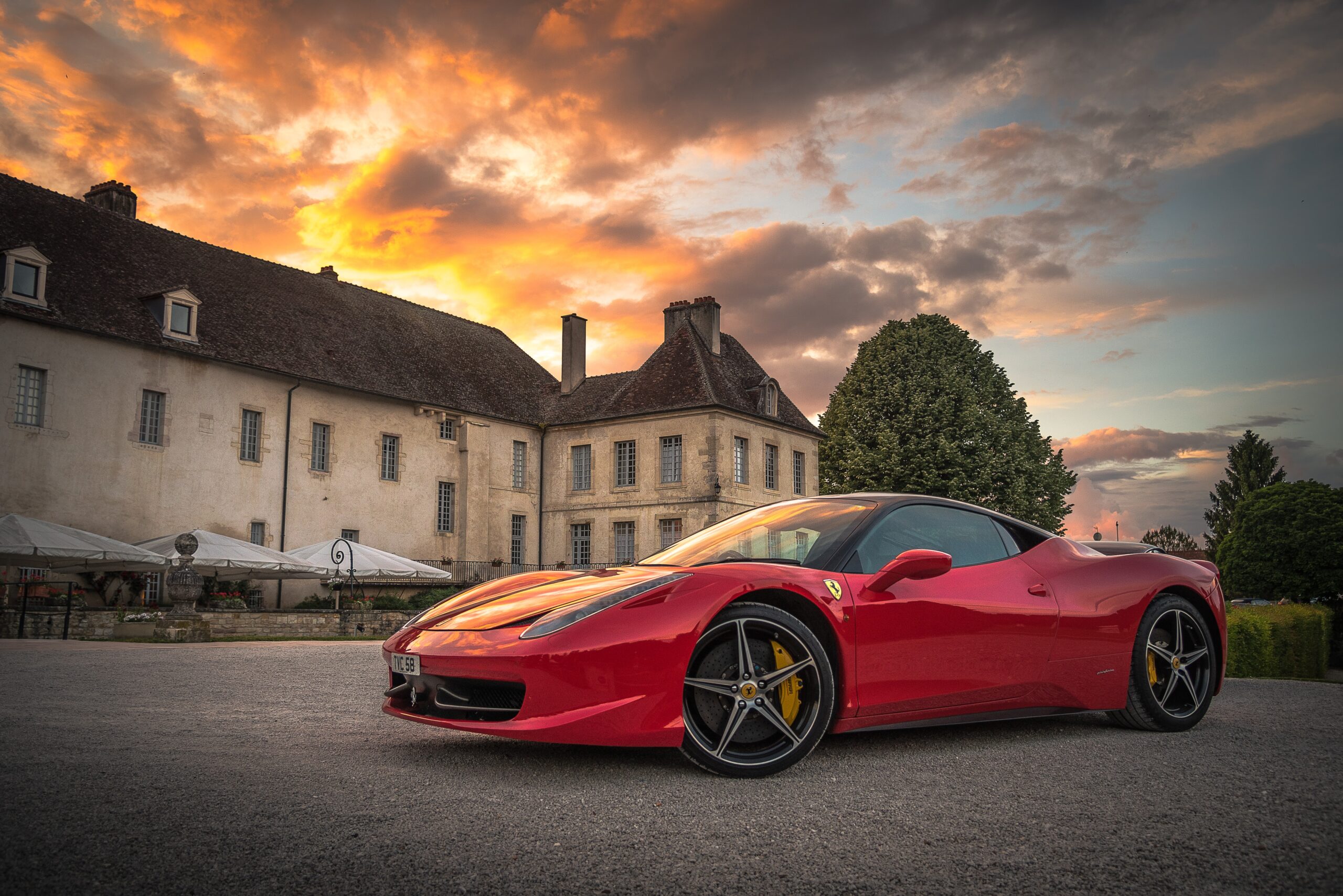 A red Ferrari viewed in profile sits on gravel in front of a sprawling European manor, below a cloudy sky with rays of sunset.